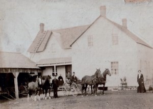 Des hommes debout à côté de chevaux et de voitures à côté d'une grande ferme blanche.